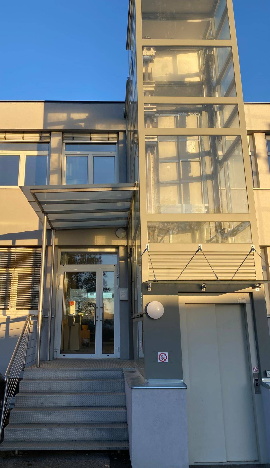 Eingangsbereich eines modernen Bürogebäudes mit Glasaufzug und Treppe.