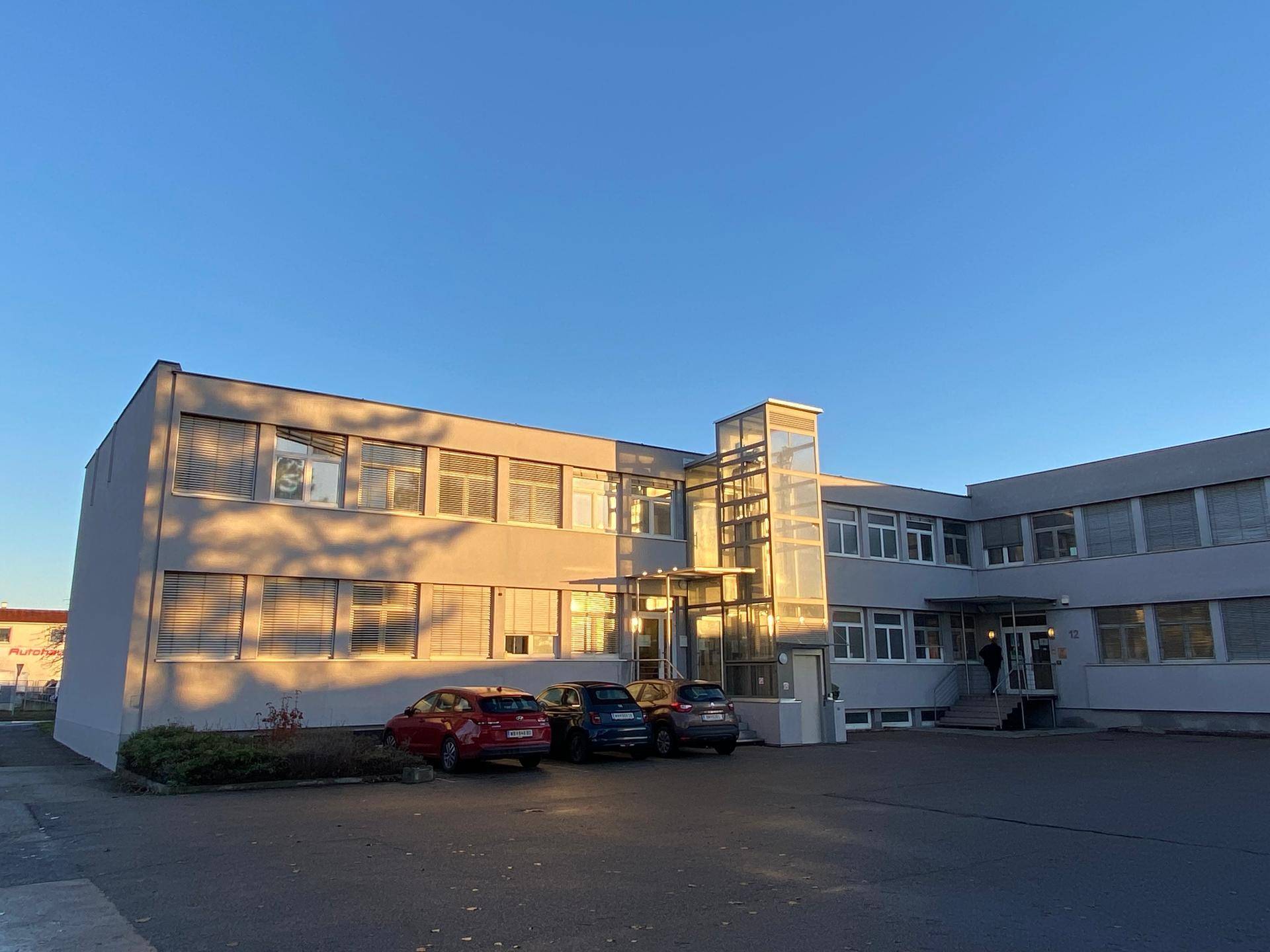 Gesamtansicht des Bürogebäudes mit Parkplätzen und einem modernen Glasaufzug bei tiefstehender Sonne.