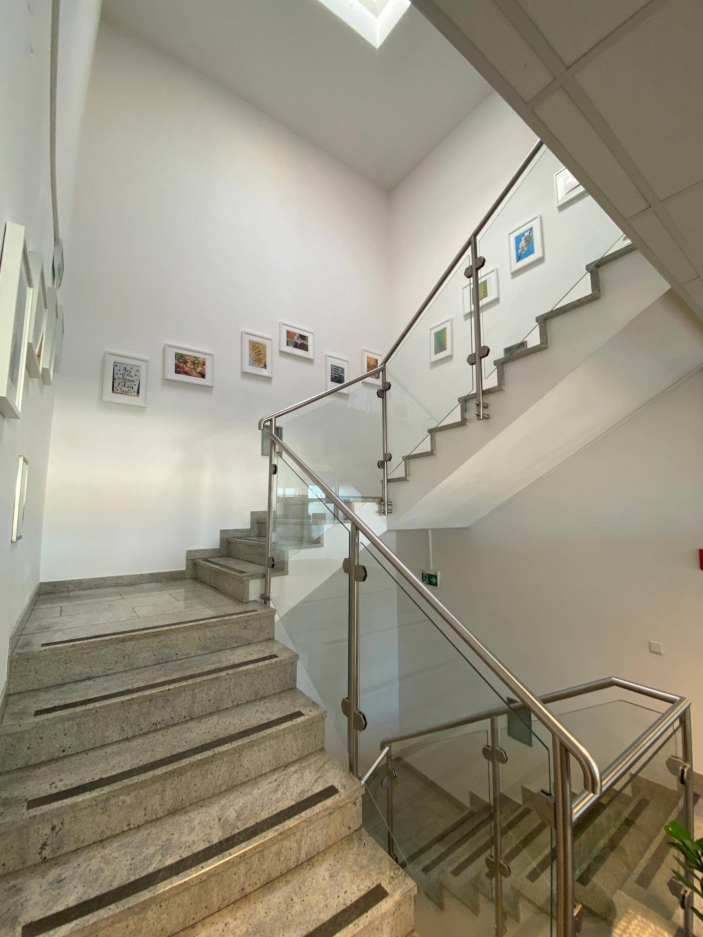 Heller Treppenaufgang mit Marmorstufen, Glasgeländer und Kunstwerken an der Wand.