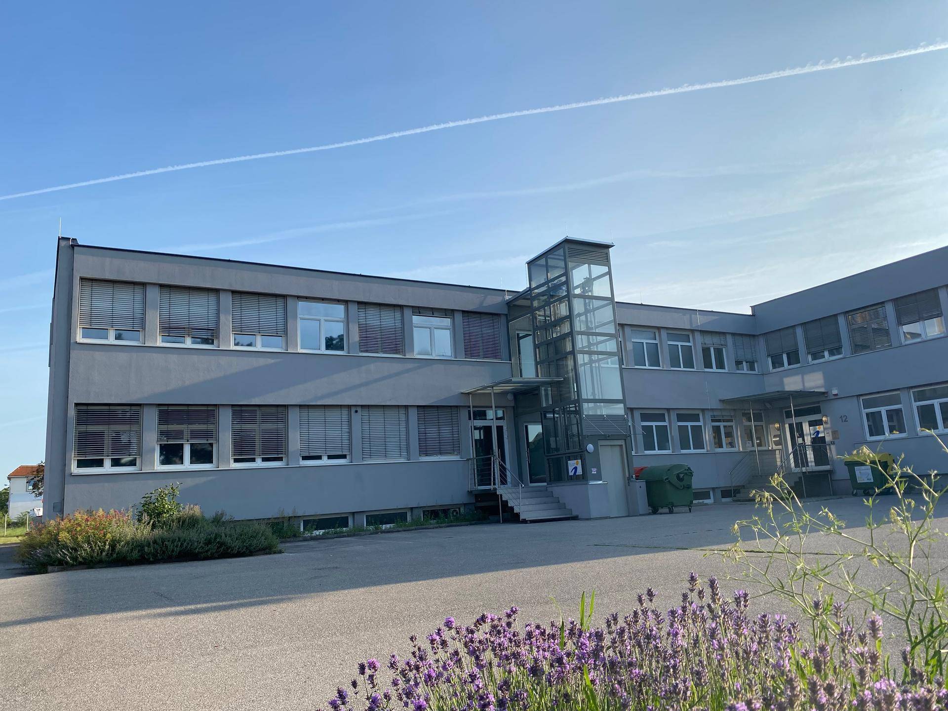 Modernes Bürogebäude mit grauer Fassade und großen Fenstern unter blauem Himmel, umgeben von Grünflächen.