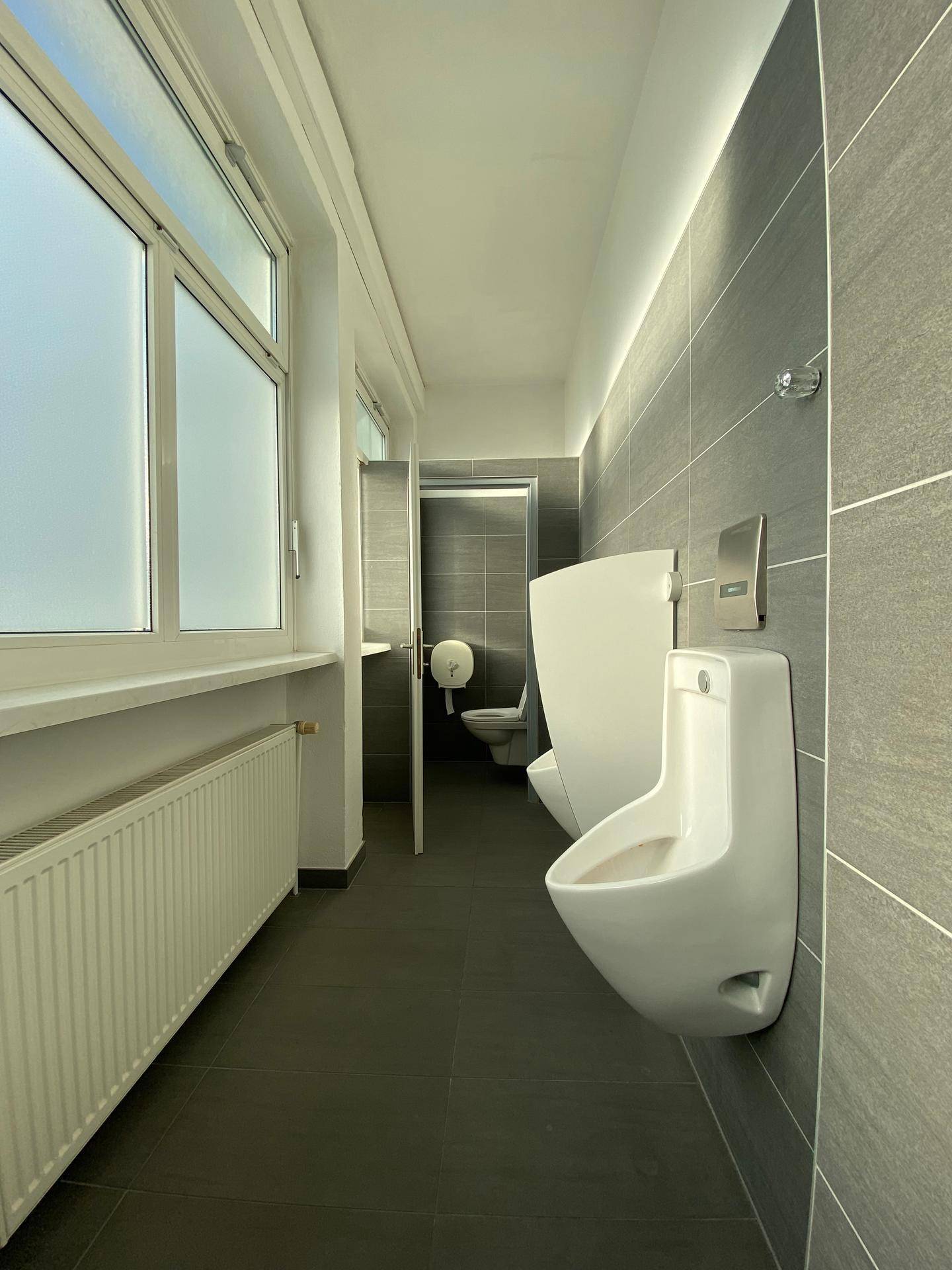 Herren-Toilette mit Urinalen, dunklen Fliesen und einem Fenster für Tageslicht.