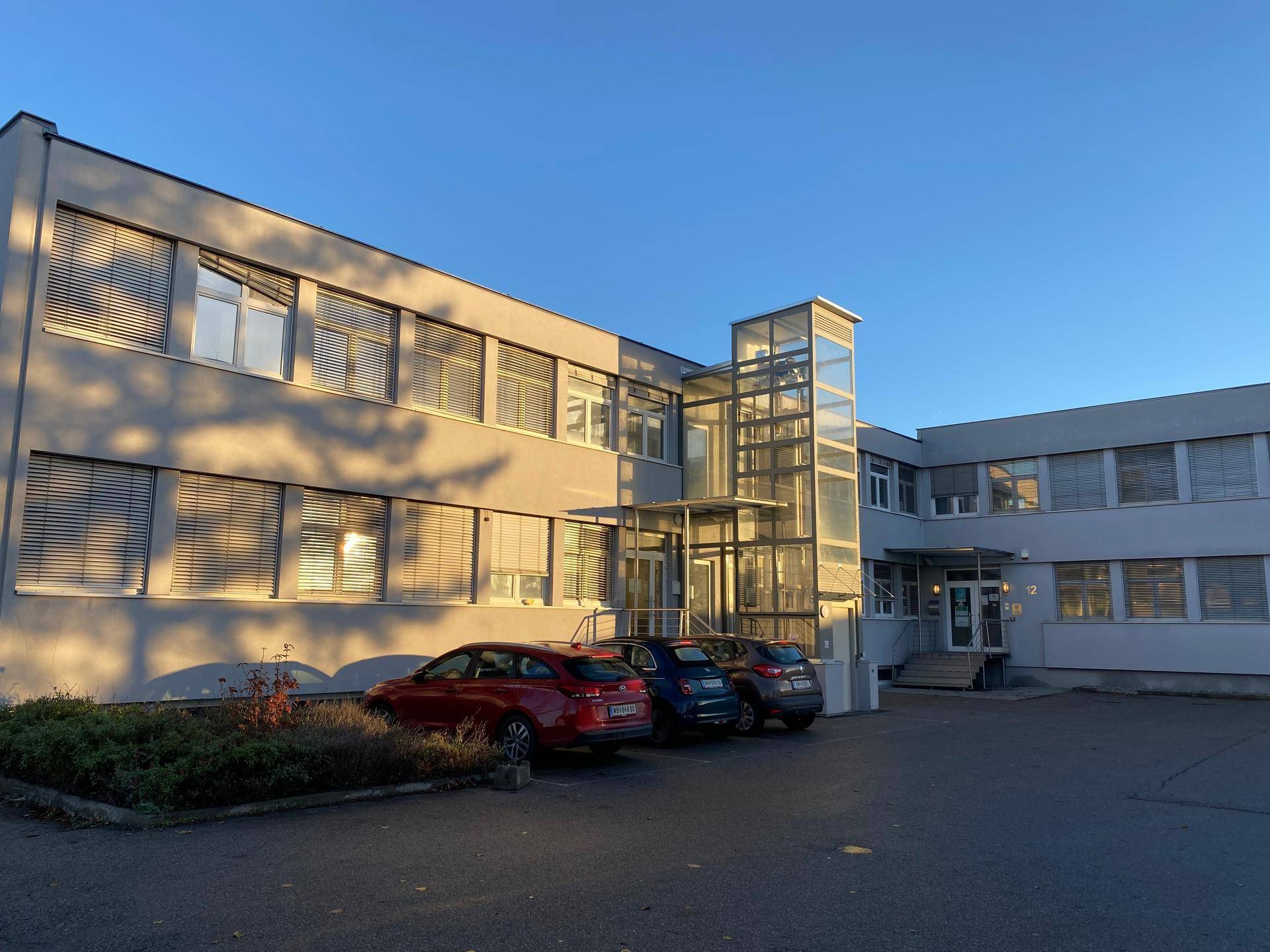 Bürogebäude mit externem Glasaufzug und geparkten Autos im warmen Abendlicht.