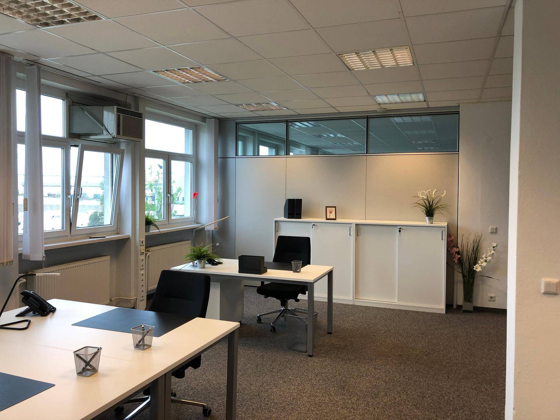 Helles und modernes Büro mit Teppichboden, großen Fenstern und funktionalen Möbeln.