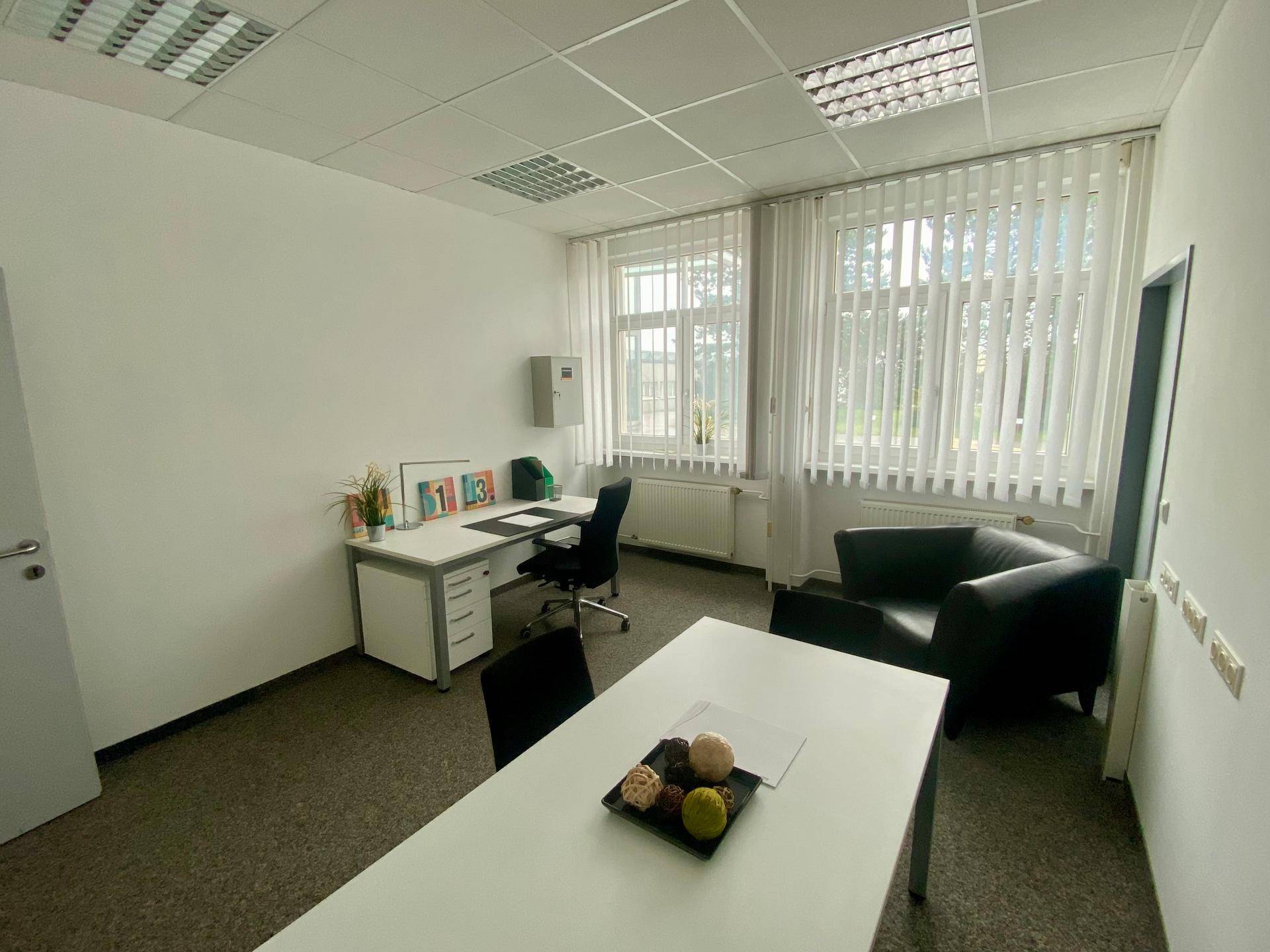 Einzelbüro mit Schreibtisch, Stuhl, Sessel und großen Fenstern mit Jalousien.