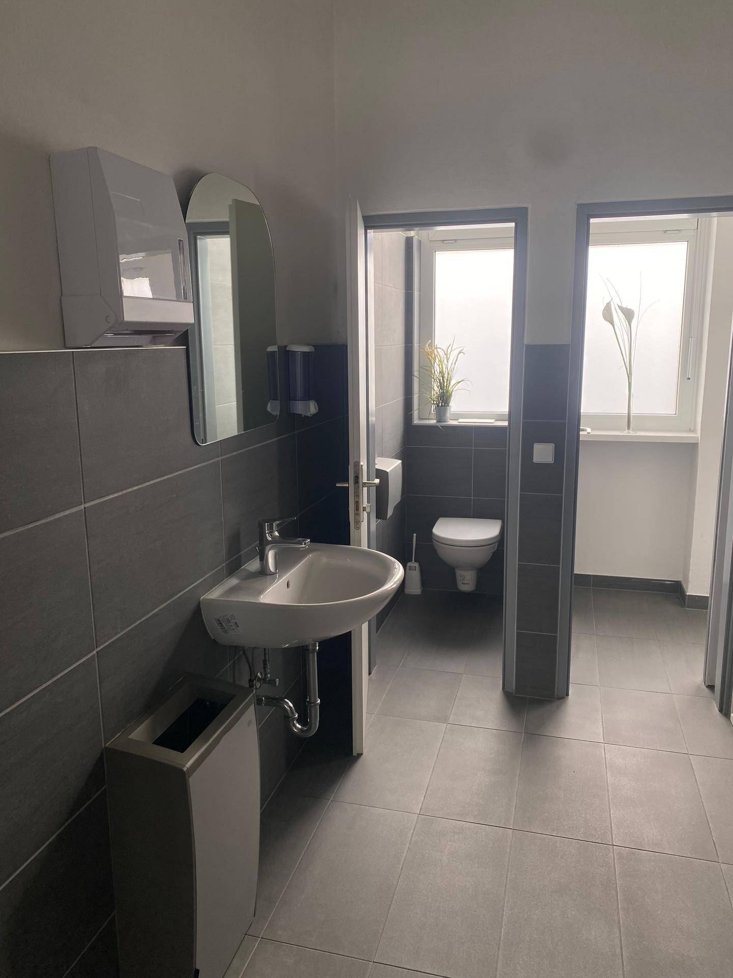 Modernes Badezimmer mit dunklen Fliesen, Waschbecken und Toilette, gut beleuchtet durch ein Fenster.