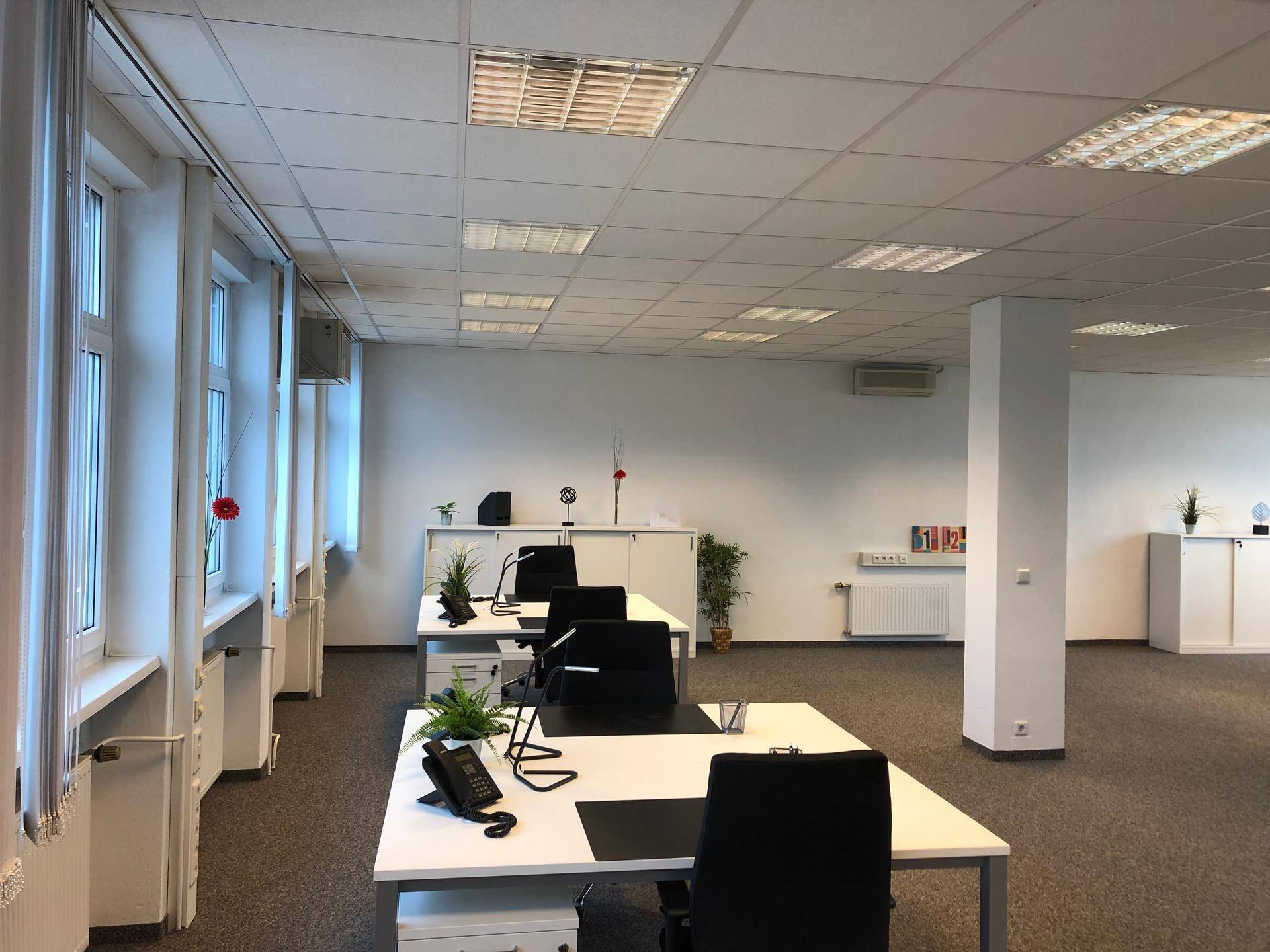 Moderner Büroraum mit mehreren Arbeitsplätzen, großen Fenstern und Teppichboden.