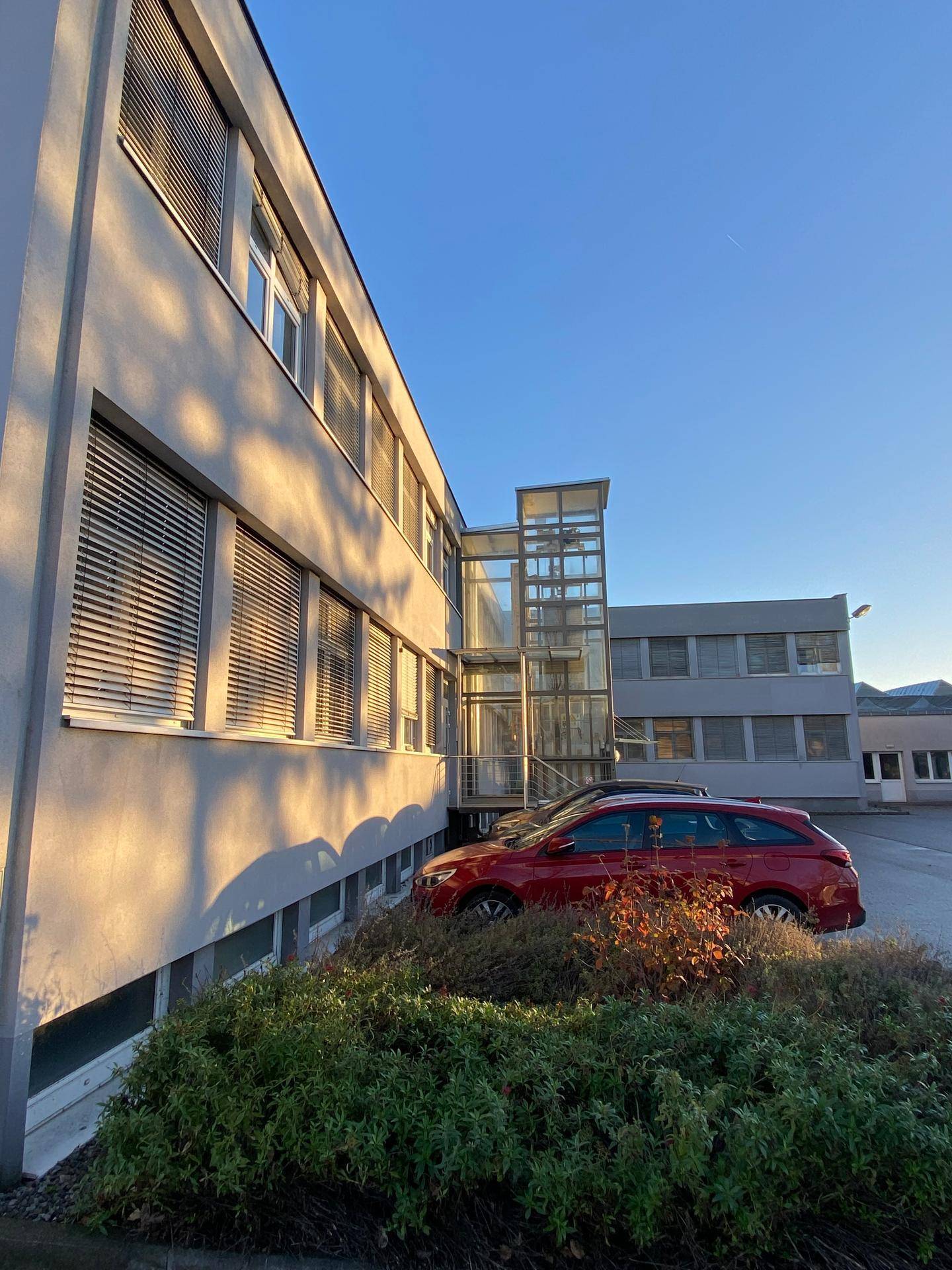 Seitenansicht des Bürogebäudes mit Glasaufzug und geparktem Auto im Sonnenlicht.