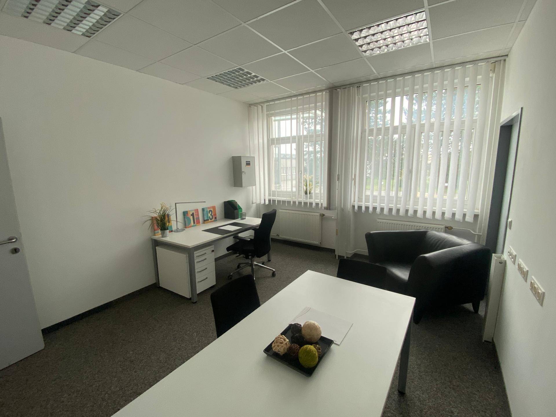 Kleines, helles Einzelbüro mit Schreibtisch, Sessel und Teppichboden, gut beleuchtet durch große Fenster.