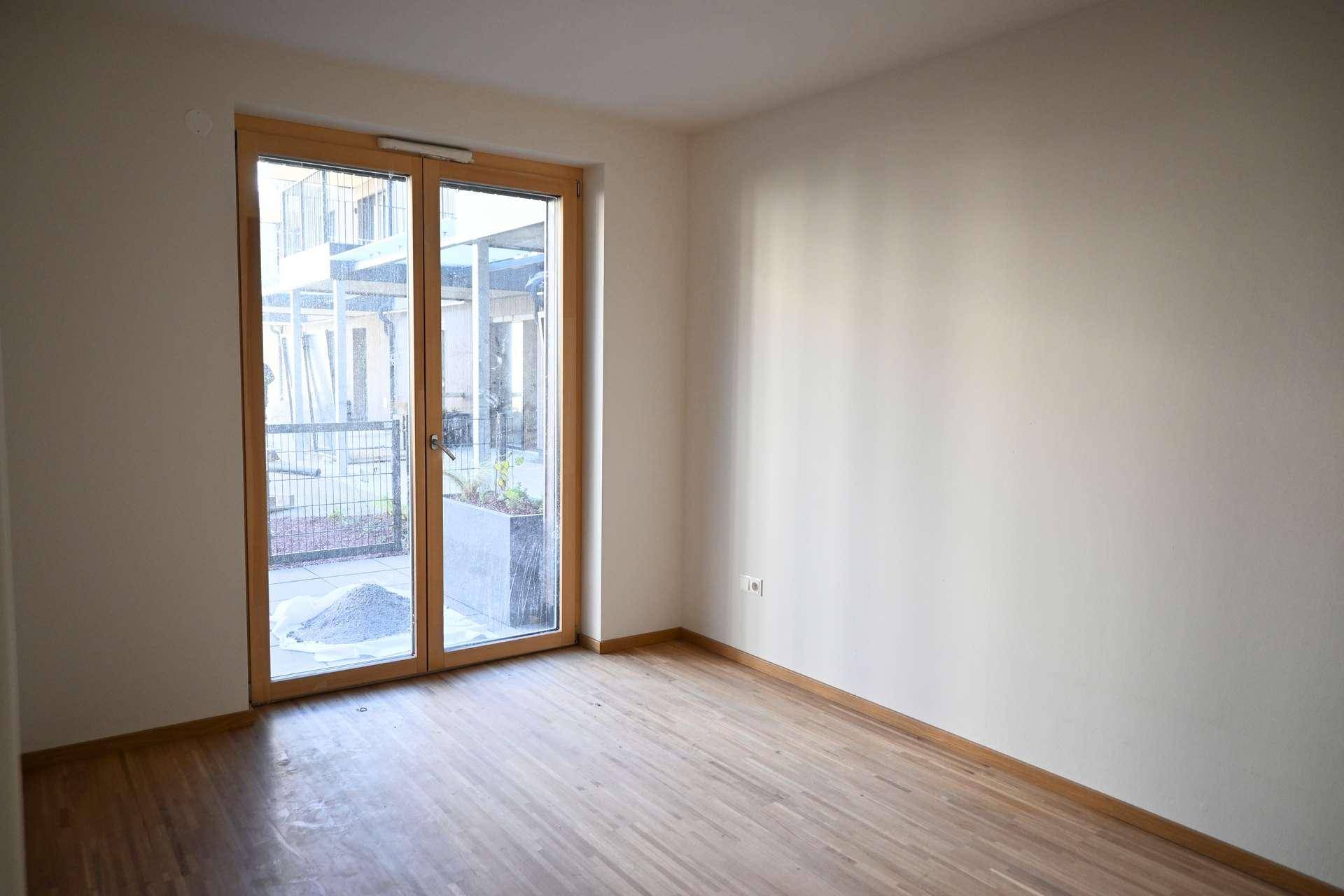 Leerer Raum mit Holzboden und einer großen Fenstertür, die viel Tageslicht hereinlässt.