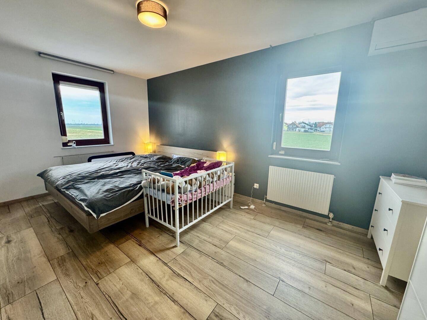 Helles Schlafzimmer mit großem Bett, Babybett und weitem Blick auf die grüne Landschaft.