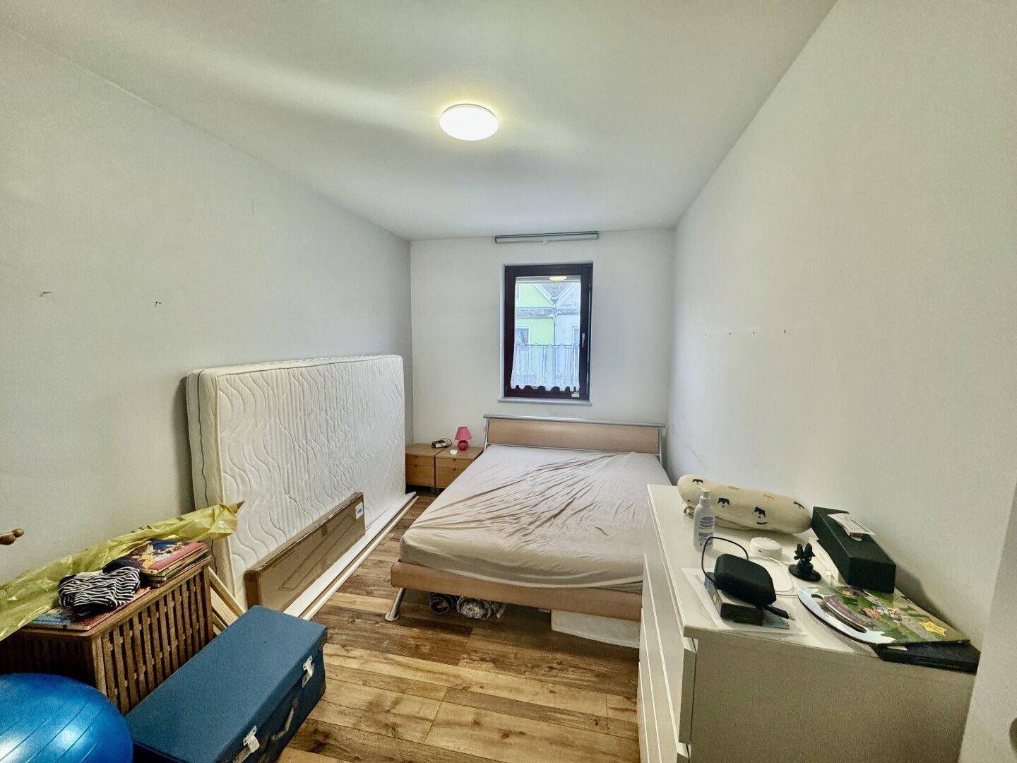 Einfaches Schlafzimmer mit Holzbett, Matratze und einem Fenster mit Blick nach draußen.