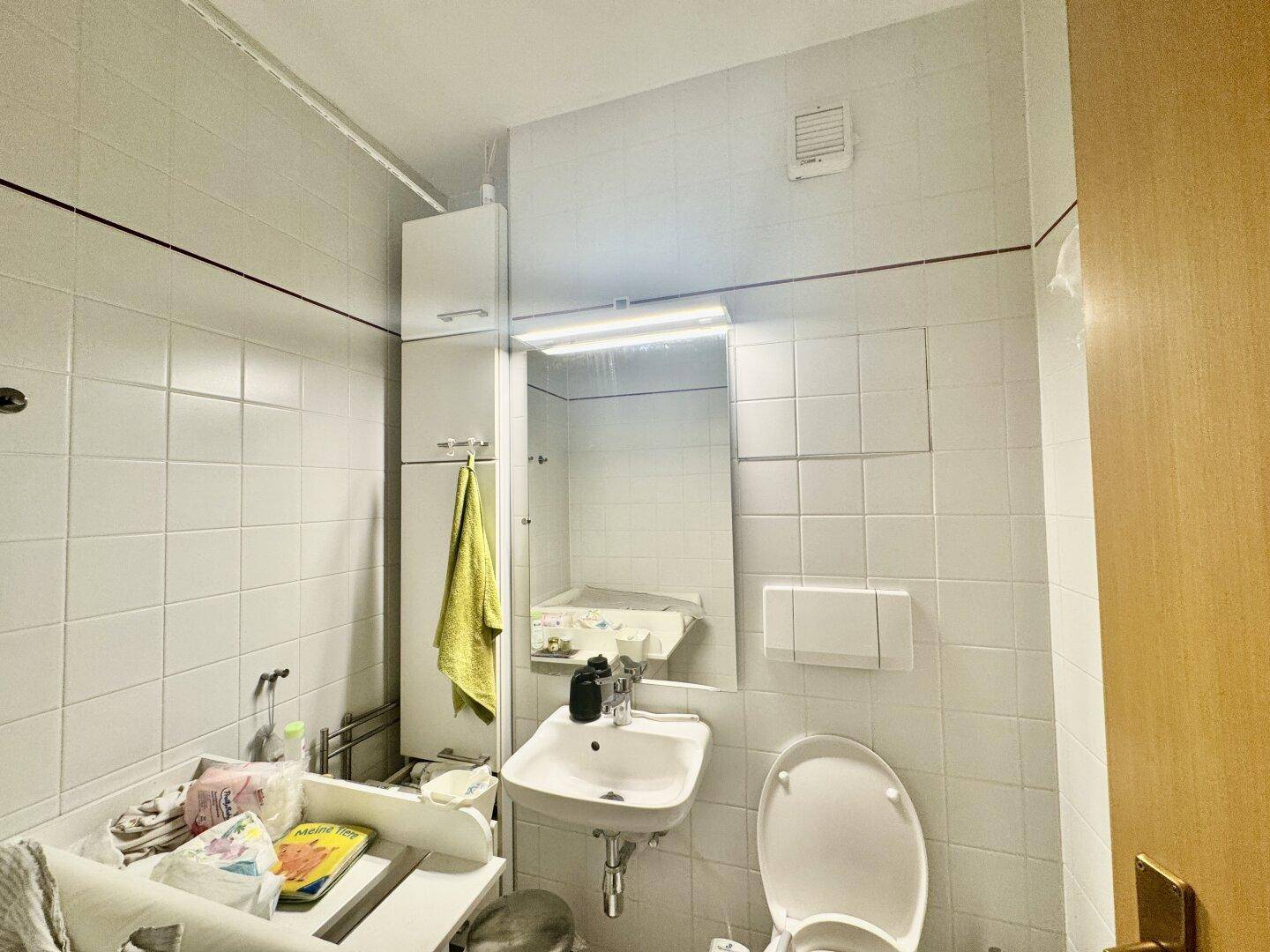 Kleines Badezimmer mit weißen Fliesen, Waschbecken, Toilette und Spiegelschrank.