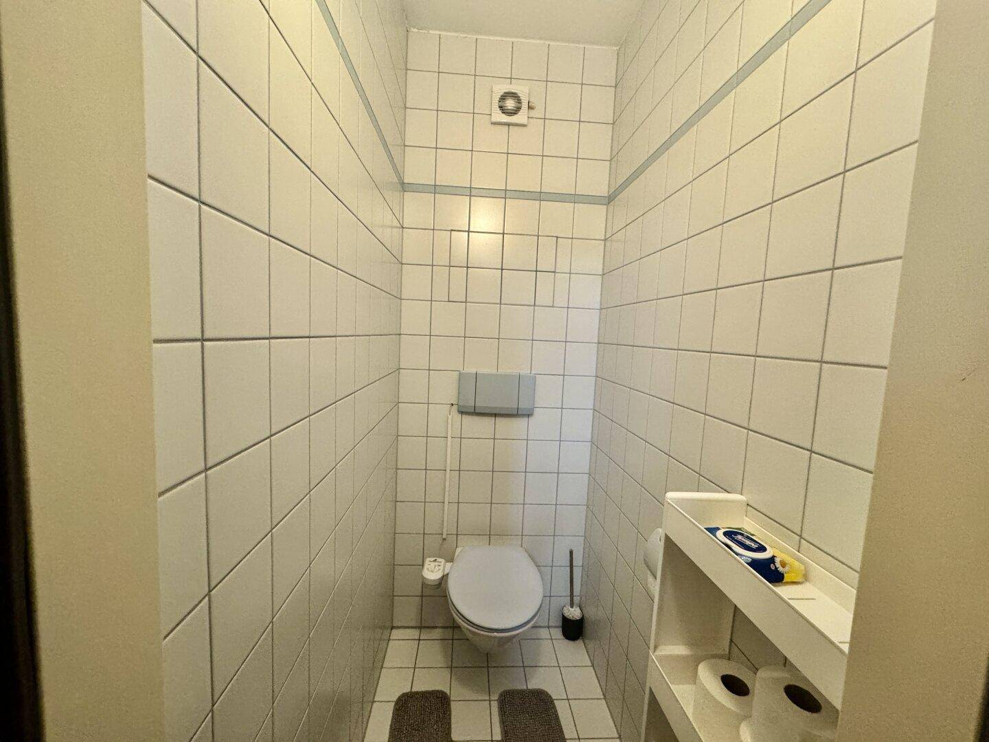 Gäste-WC mit weißen Fliesen, wandhängender Toilette und Lüftungsanlage.