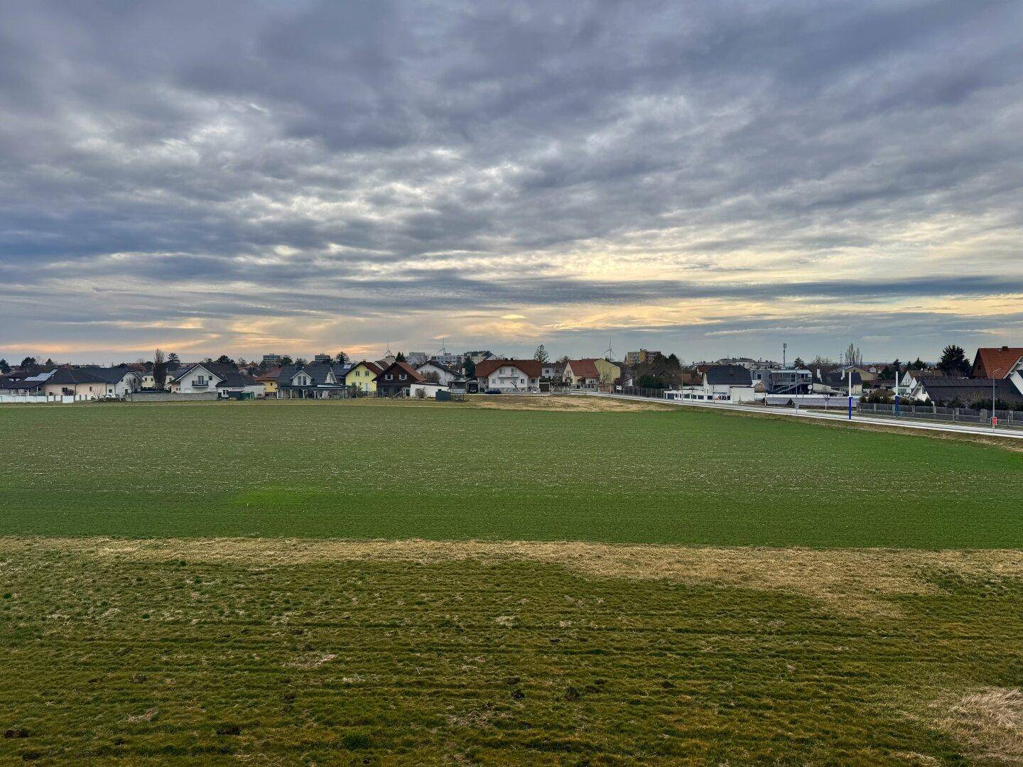 Weitläufiger Blick über grüne Felder auf eine ländliche Wohnsiedlung unter bewölktem Himmel.