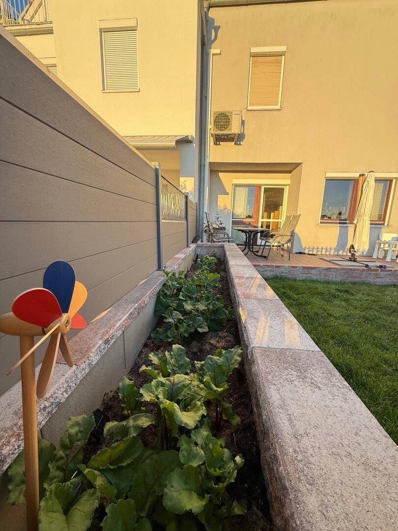 Erhöhtes Blumenbeet mit Pflanzen und einem Windrad, neben der Hausfassade.