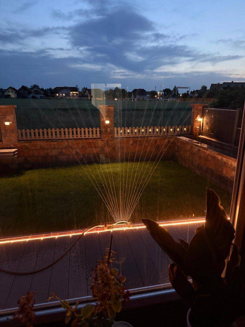 Garten bei Dämmerung mit Rasensprenger und stimmungsvoller Außenbeleuchtung.