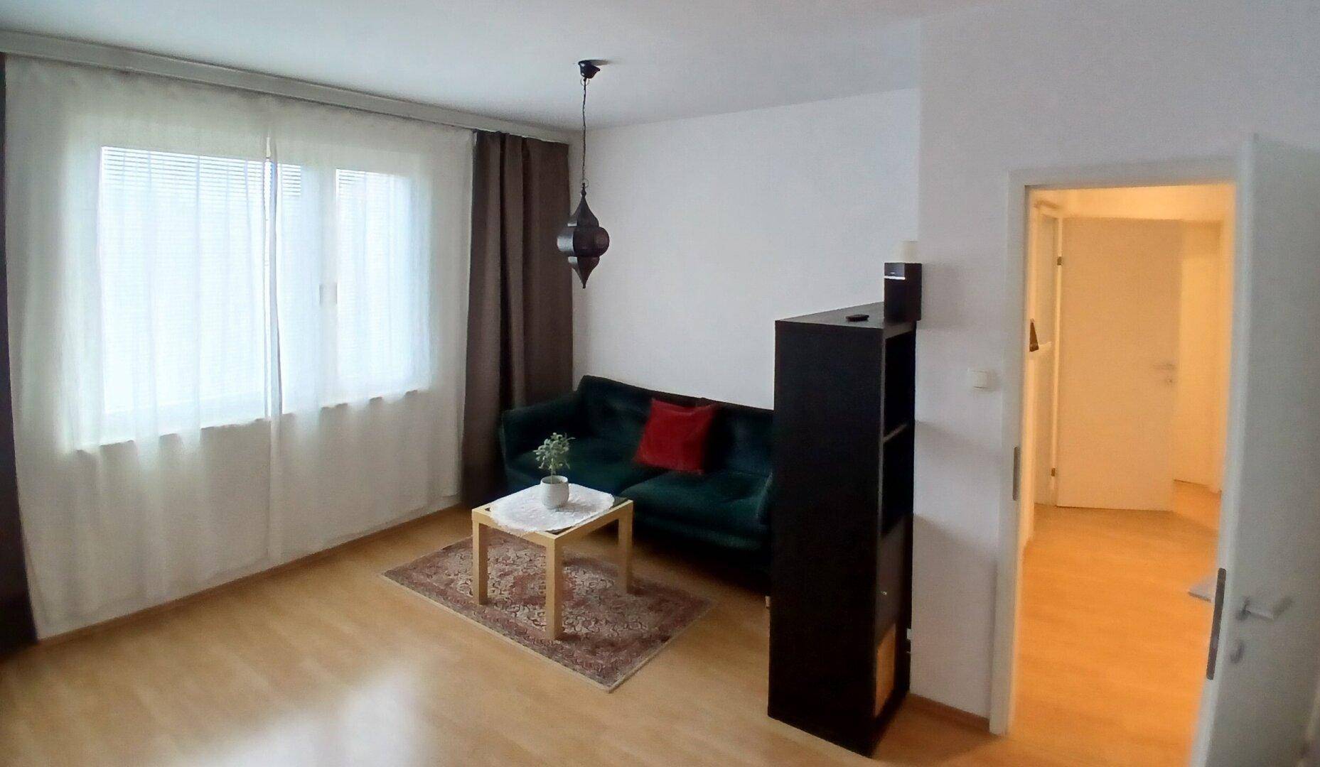 Gemütliches Wohnzimmer mit grünem Sofa, Couchtisch und großem Fenster mit Vorhängen.