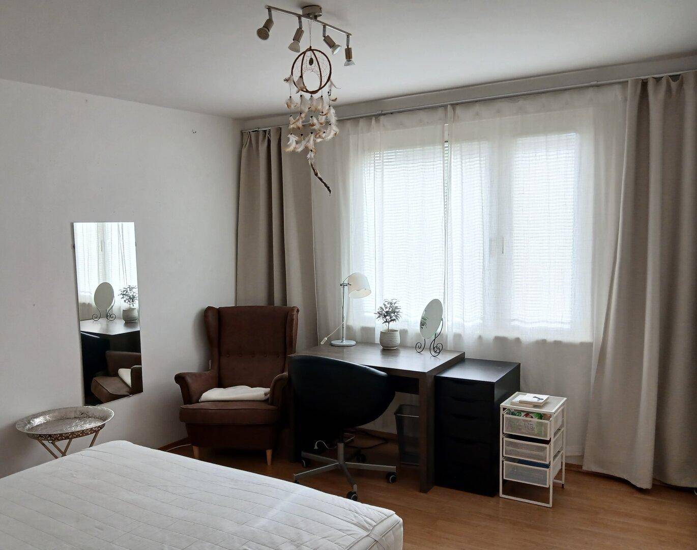 Helles Schlafzimmer mit Bett, Schreibtisch und einem großen Fenster, das viel Tageslicht hereinlässt.