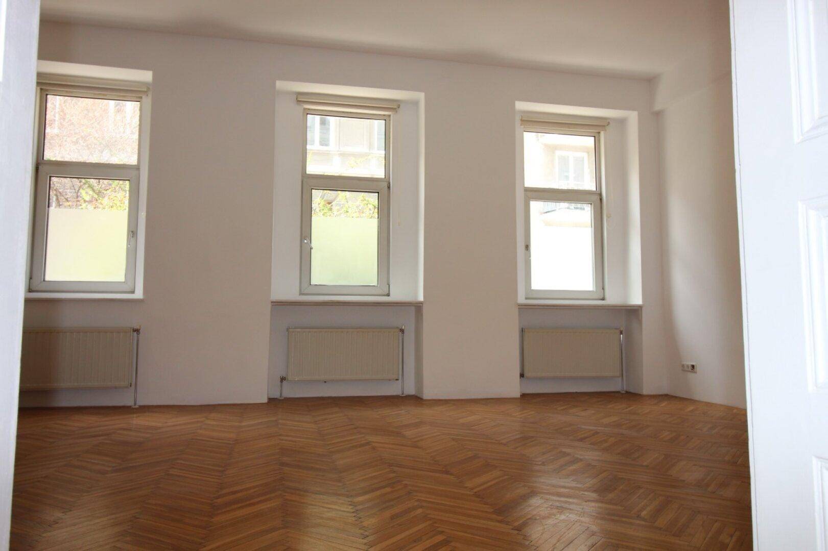 Geräumiges Zimmer mit klassischem Fischgrätparkett und drei großen Fenstern für viel Tageslicht.