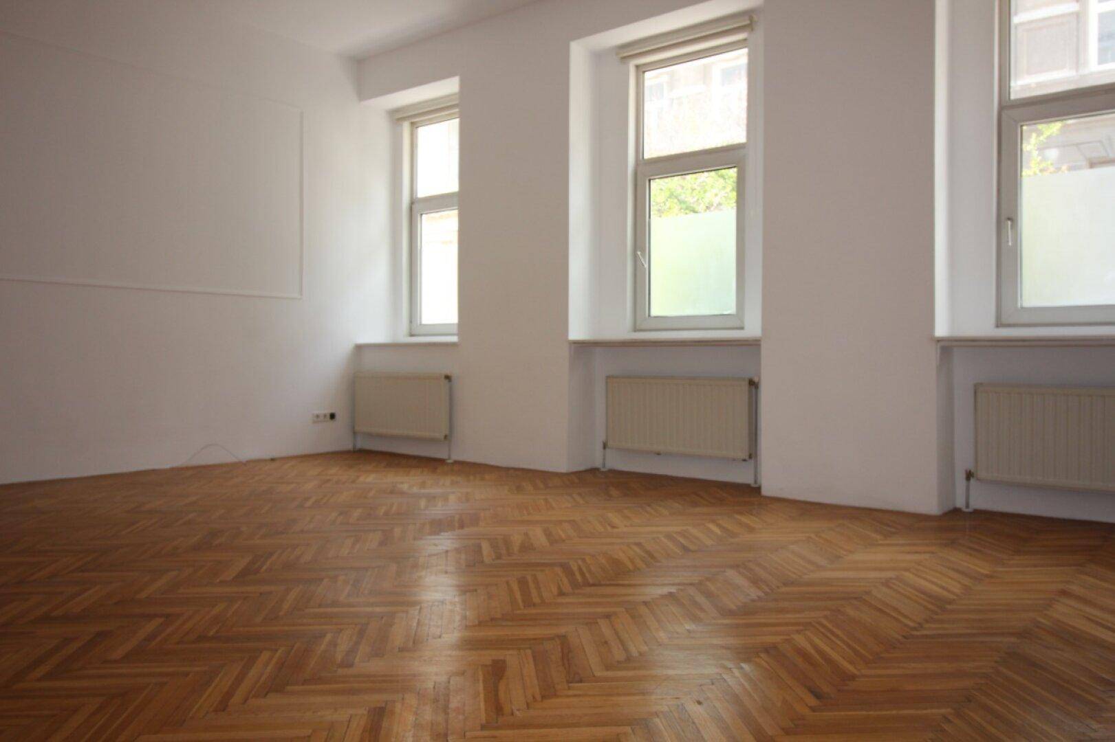 Lichtdurchfluteter Raum mit elegantem Fischgrätparkett und hohen Fenstern, ideal für Wohnzwecke.