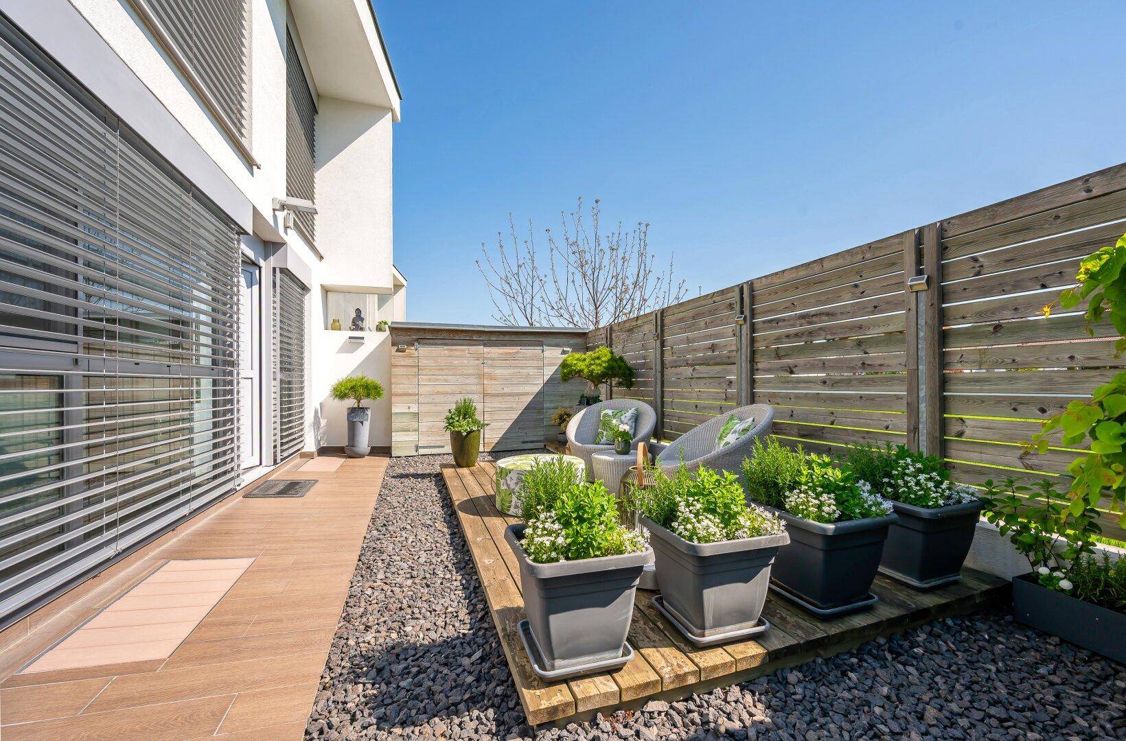 Kleine, gemütliche Terrasse mit Kiesbett, Holzdeck und Topfpflanzen, umgeben von einem Holzzaun.