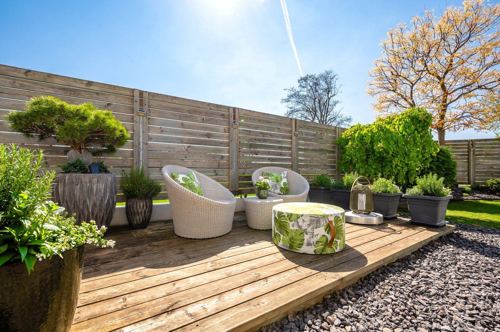 Sonnige Terrasse mit modernen Korbmöbeln, Holzzaun und üppiger Bepflanzung im Hintergrund.