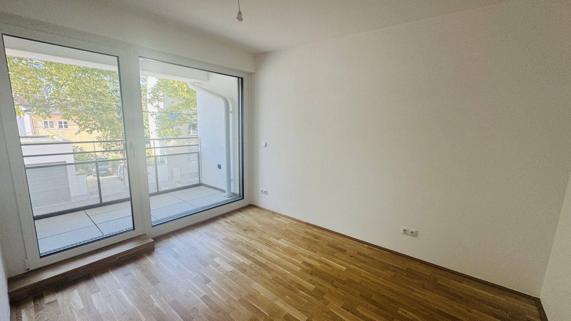 Helles Schlafzimmer mit Parkettboden und großem Fenster mit Balkonzugang.