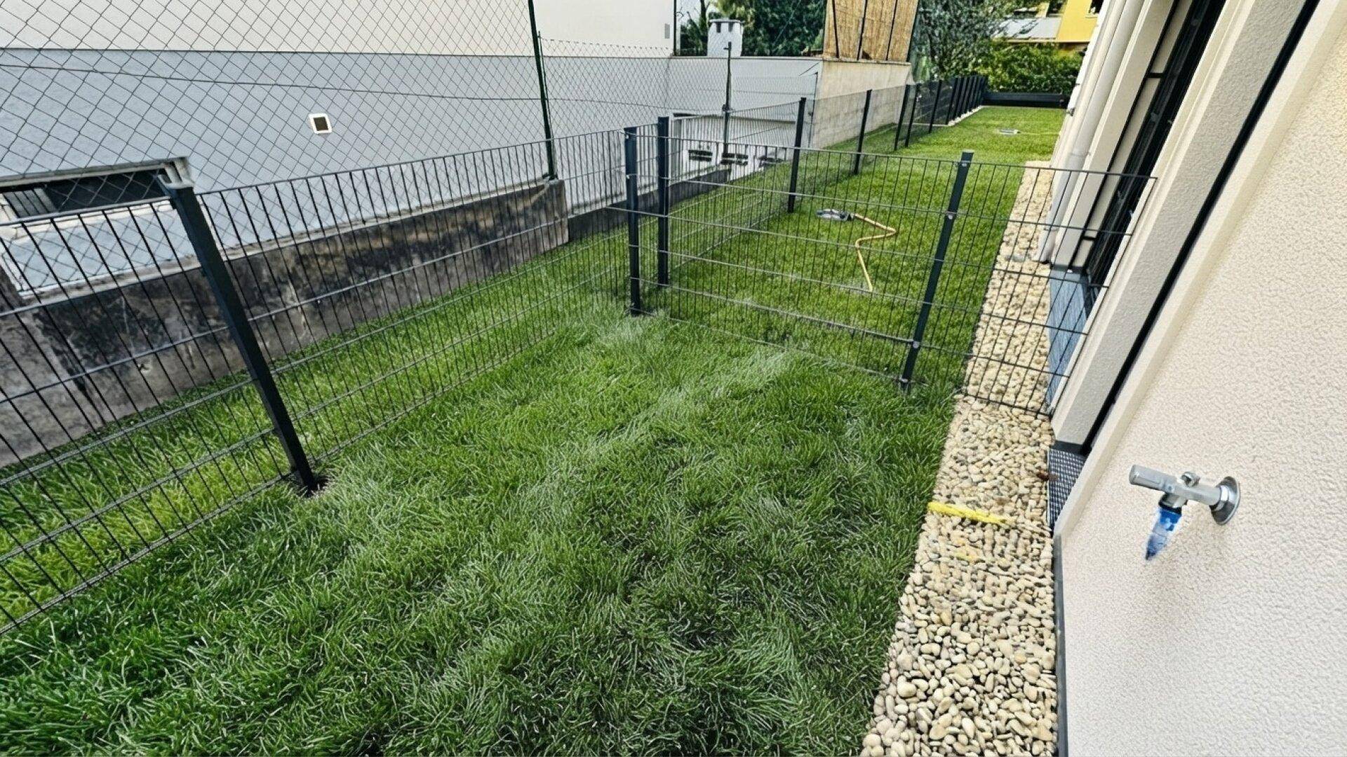 Eingezäunter Gartenbereich mit frischem Rasen und Kiesstreifen am Gebäude.