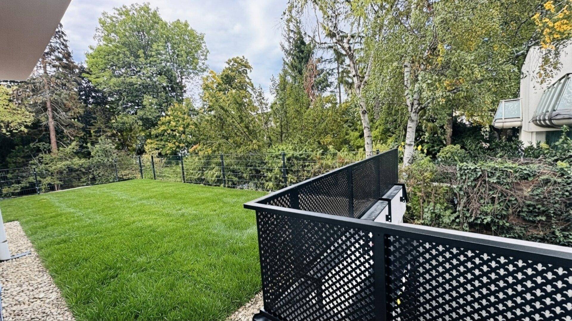 Gepflegter Gartenbereich mit Rasenfläche, Zaun und altem Baumbestand im Hintergrund.