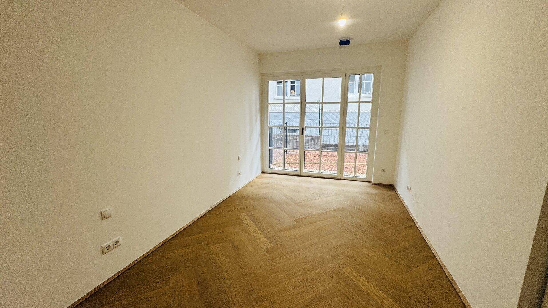 Helles Zimmer mit Fischgrätparkett und großem Fenstertür-Element zum Außenbereich.