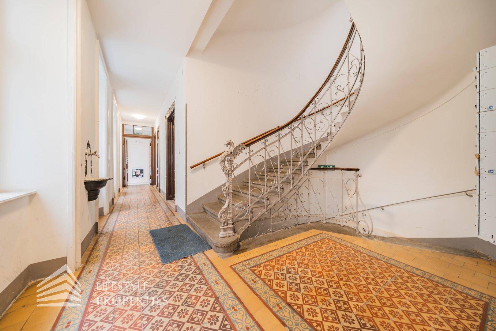 Historisches Treppenhaus mit elegantem schmiedeeisernem Geländer, Steinstufen und gemustertem Fliesenboden.