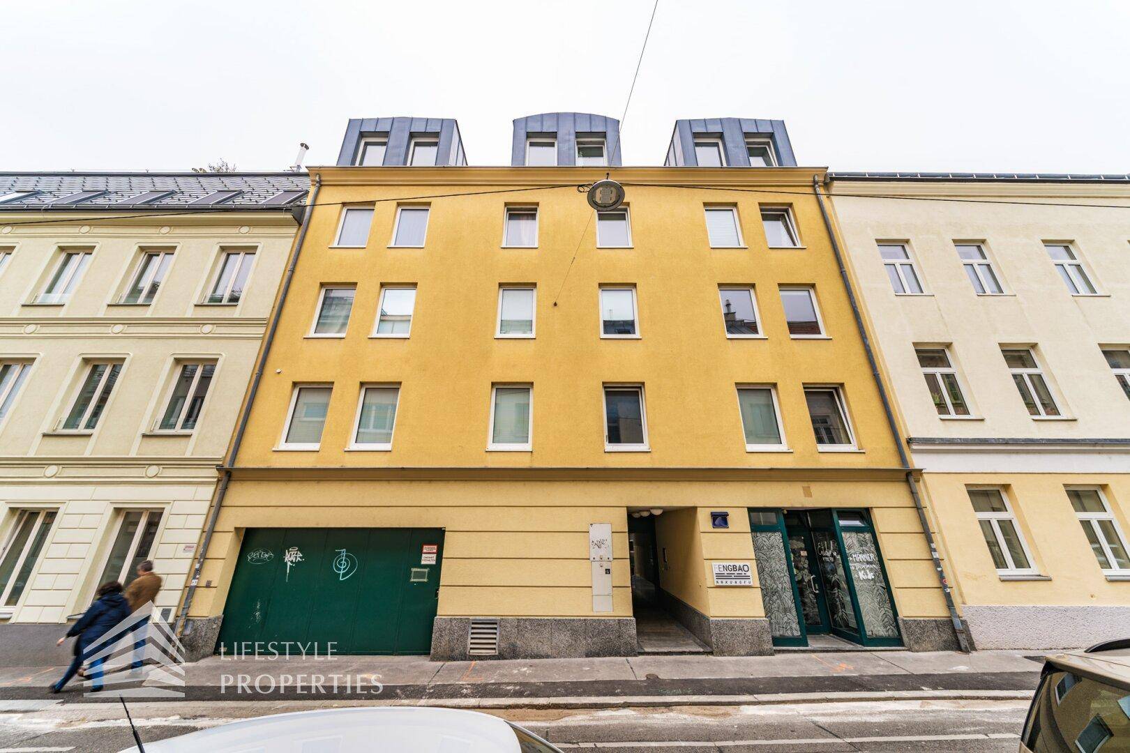 Gesamtansicht der Wohnhausfassade mit Tiefgarageneinfahrt und Geschäften im Erdgeschoss.