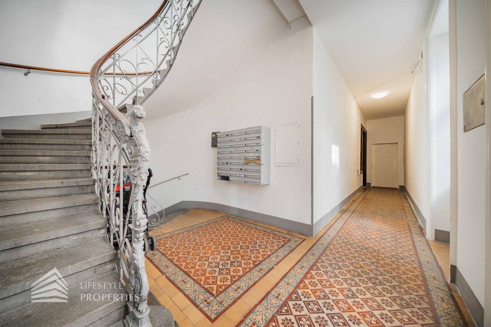 Historisches Treppenhaus mit kunstvollem Geländer, Steinstufen und gemustertem Fliesenboden im Eingangsbereich.
