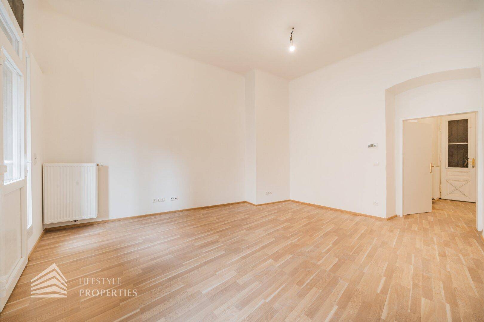 Geräumiger, heller Raum mit Parkettboden und Zugang zu einem weiteren Bereich durch einen Torbogen.