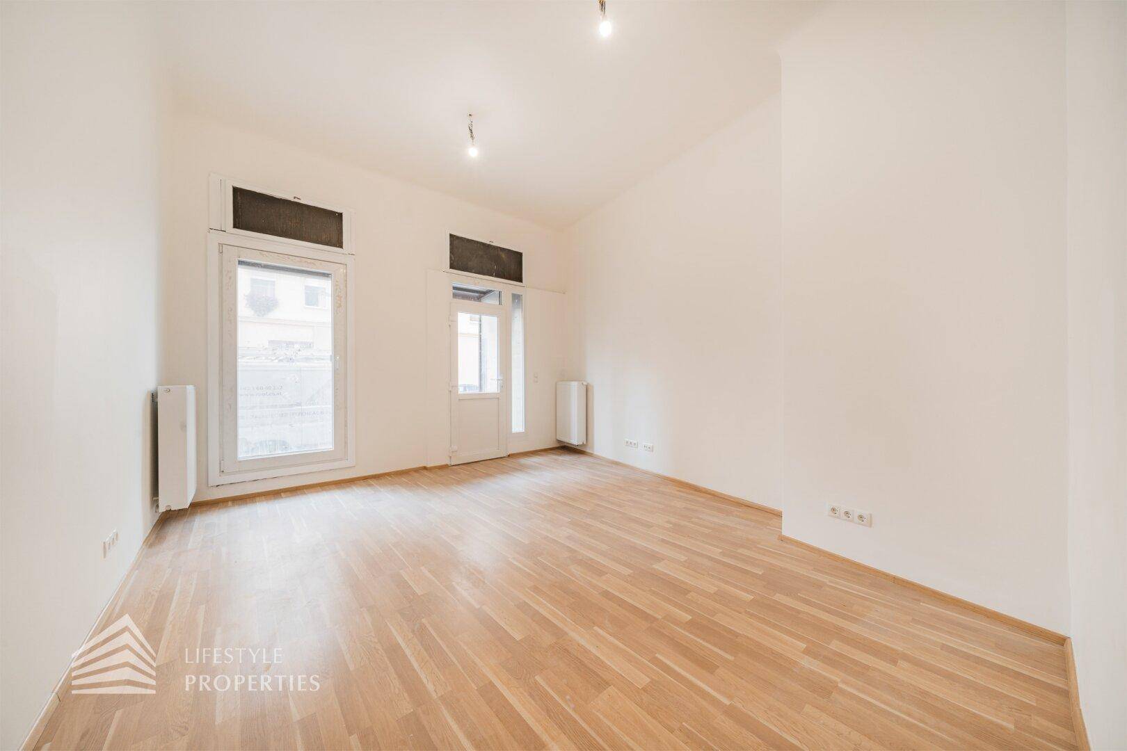 Heller, leerer Raum mit Holzboden und zwei Fenstertüren, die viel Tageslicht hereinlassen.