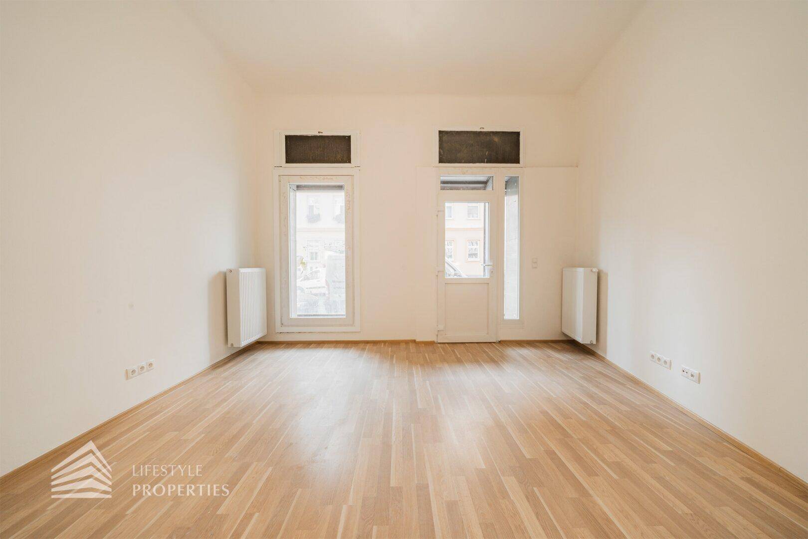 Leerer Raum mit hellem Holzboden und zwei großen Fenstertüren, die den Raum mit Licht füllen.