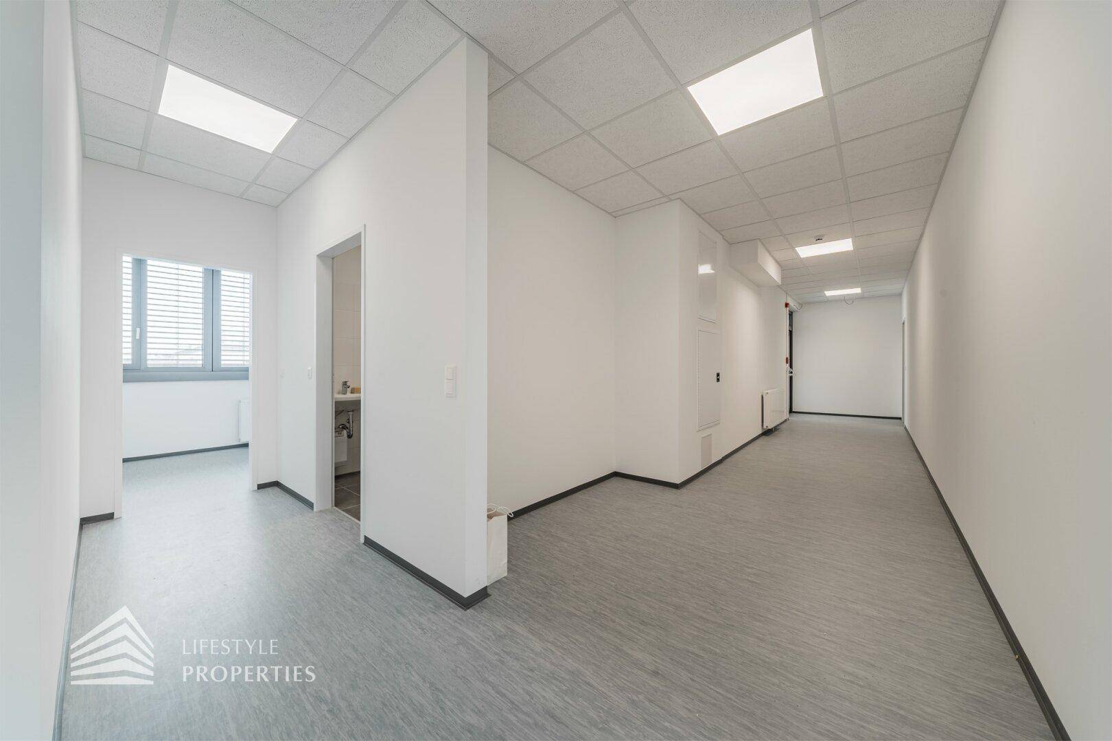 Langer, heller Flur in einem Bürogebäude mit abgehängter Decke und Zugang zu verschiedenen Räumen.