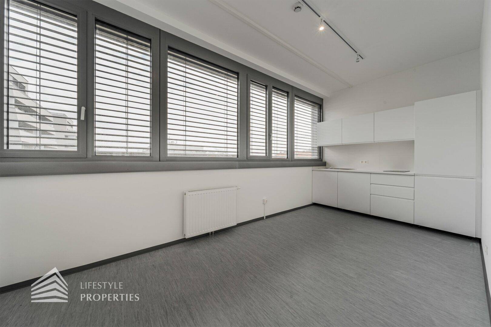 Büroraum mit integrierter, weißer Küchenzeile und großen Fenstern mit Jalousien für flexible Lichtregulierung.