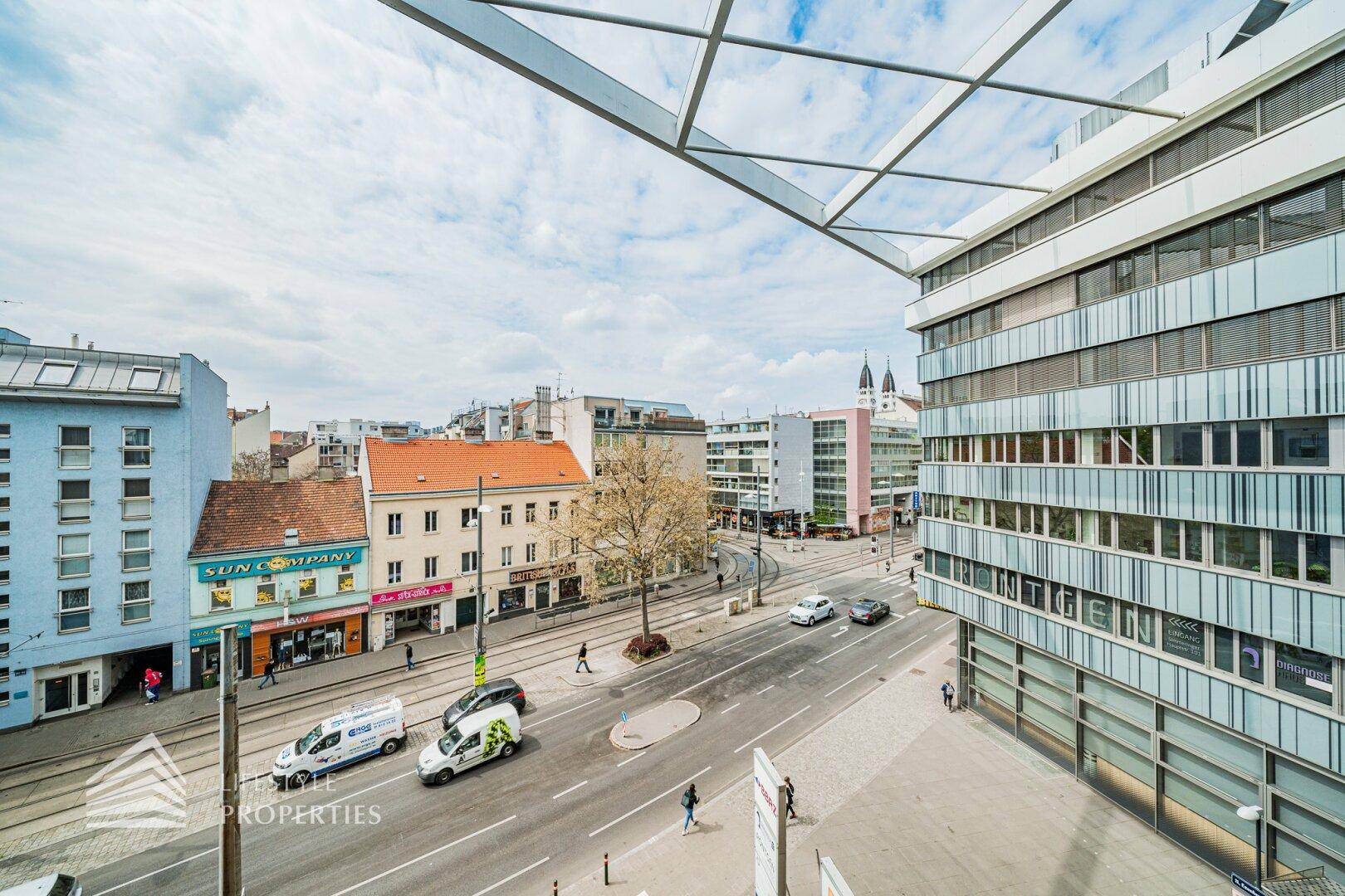 Panoramablick auf eine belebte Stadtstraße mit Gebäuden, Fahrzeugen und Fußgängern unter bewölktem Himmel.