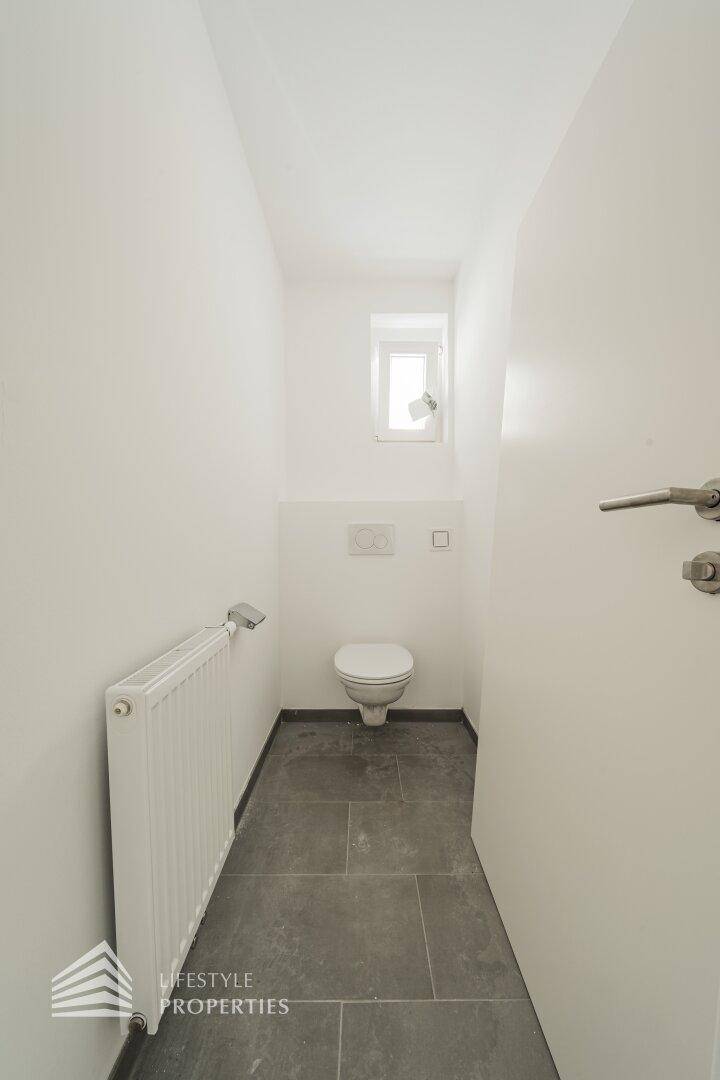 Separates WC mit wandhängender Toilette, Fenster und Fliesenboden.