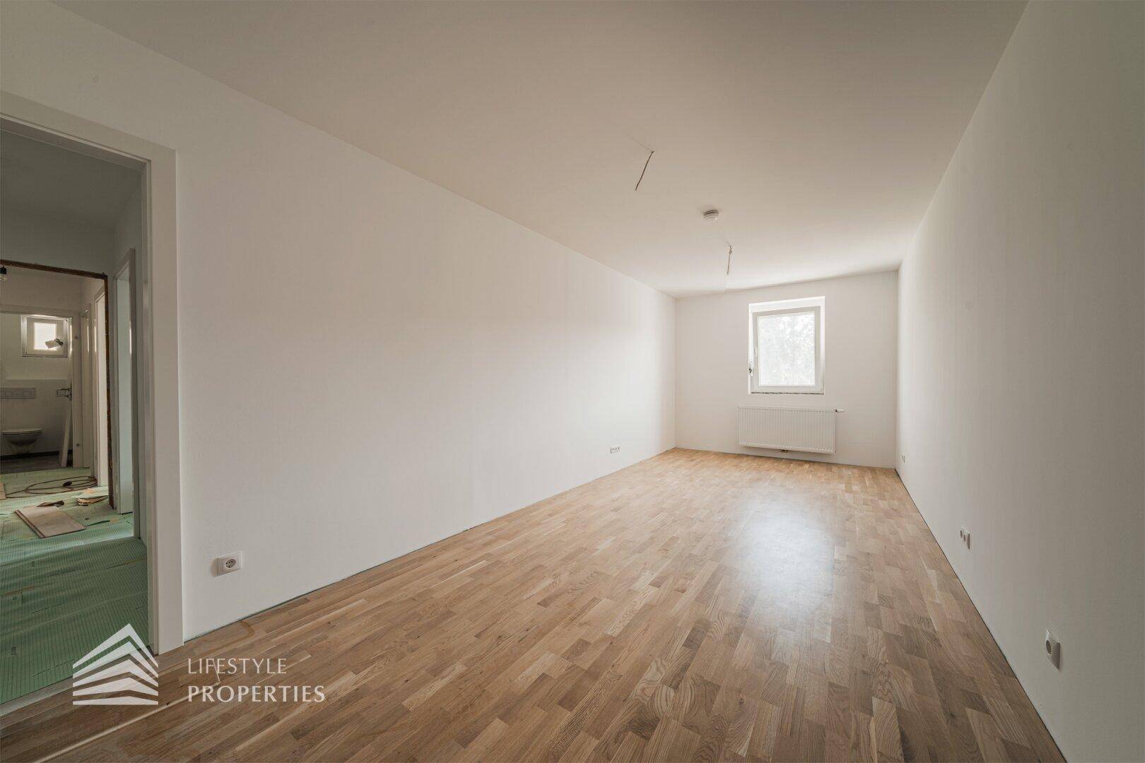 Geräumiges, helles Zimmer mit Holzboden und Blick auf den angrenzenden Badbereich.