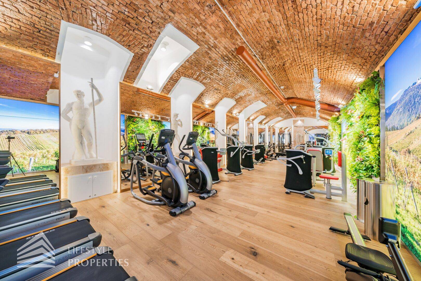 Fitnessraum mit modernen Trainingsgeräten, Holzboden und beeindruckendem Ziegelgewölbe an der Decke.