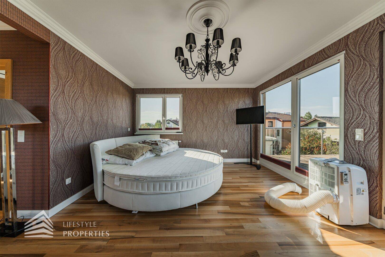 Geräumiges Schlafzimmer mit rundem Bett, großen Fenstern, Terrassenzugang und mobiler Klimaanlage.