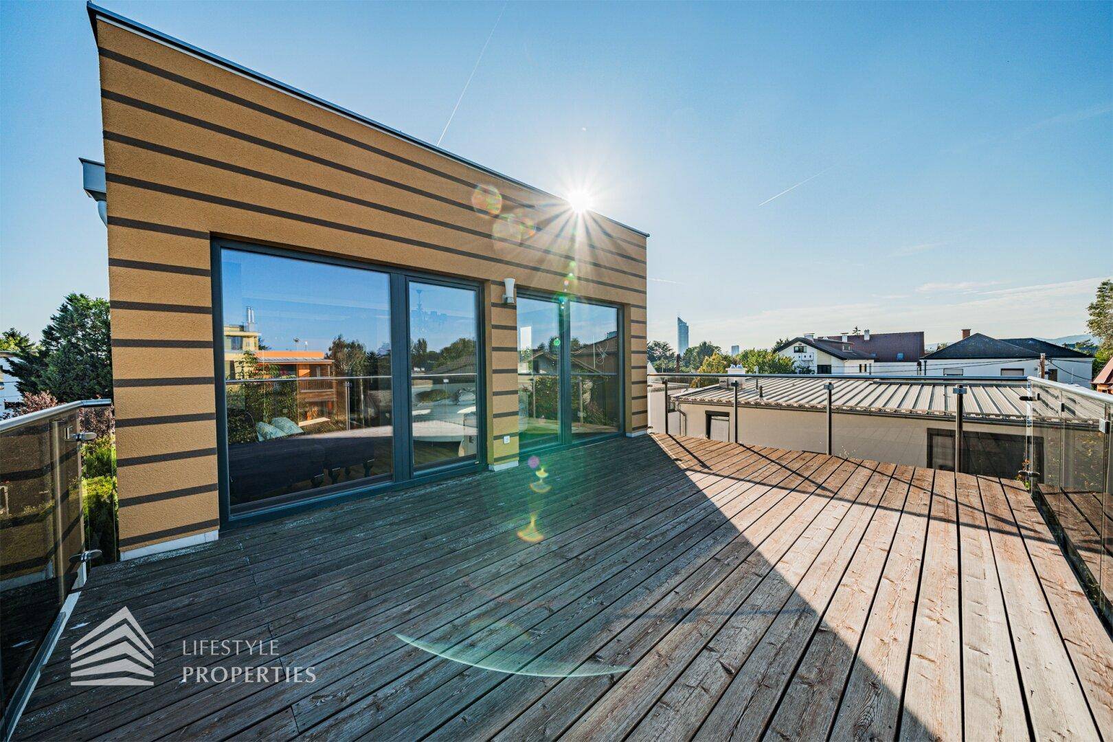 Moderne Gebäudefassade mit großen Fenstern und einer weitläufigen Holzterrasse unter blauem Himmel.