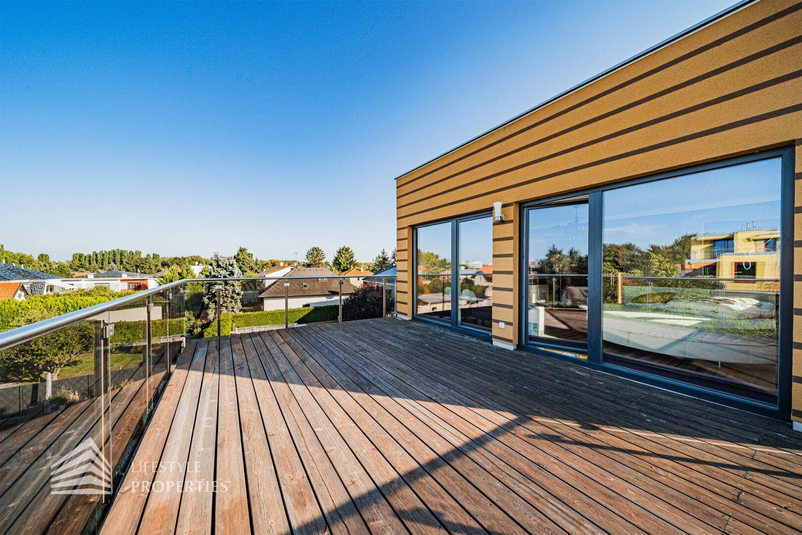 Große Holzterrasse mit Glasgeländer und weitem Blick über die Nachbarschaft unter blauem Himmel.
