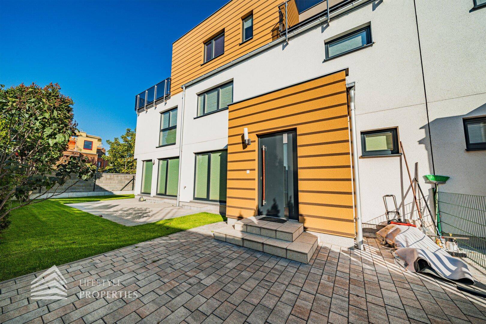 Rückseite des modernen Hauses mit gepflasterter Terrasse und direktem Zugang zum Garten.