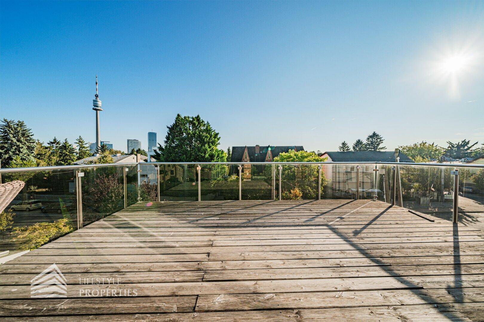 Große Holzterrasse mit Glasgeländer und weitem Blick über die Stadt und den Donauturm.