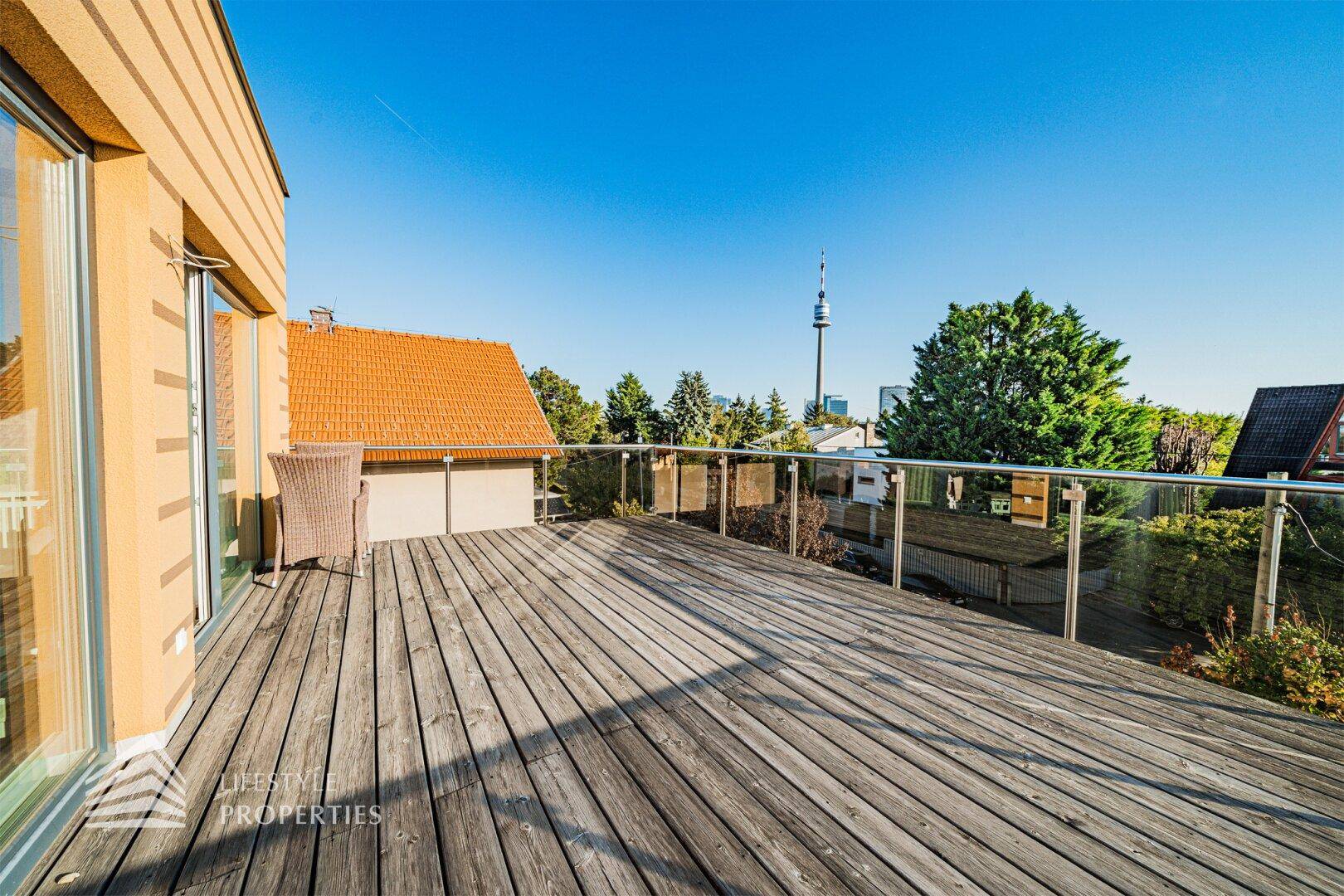 Geräumige Holzterrasse mit Zugang zum Gebäude und Blick auf die Umgebung und den Donauturm.