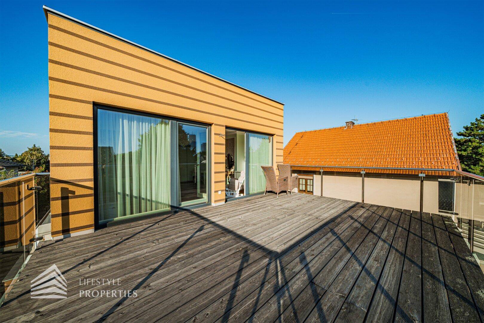 Sonnige Holzterrasse mit großen Fenstern und modernem Gebäude im Hintergrund, ideal zum Entspannen.