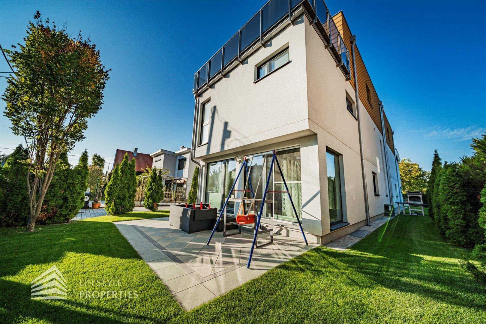 Moderne Hausfassade mit großzügiger Rasenfläche, Terrasse und Kinderspielbereich im Freien.